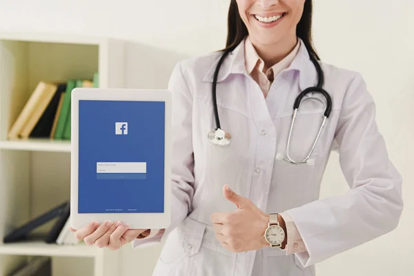 Cropped view of smiling doctor showing thumb up and presenting tablet with facebook app — Stock Photo
