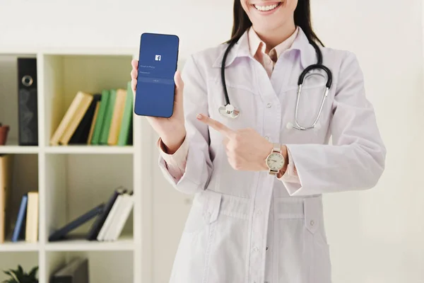 Cropped view of doctor with stethoscope pointing at smartphone with facebook app — Stock Photo