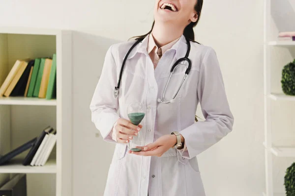 Vista cortada de médico rindo em casaco branco com estetoscópio segurando relógio de areia — Fotografia de Stock