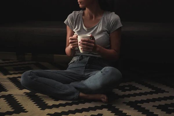 Vista recortada de la mujer triste con taza de café sentado en el suelo en casa - foto de stock