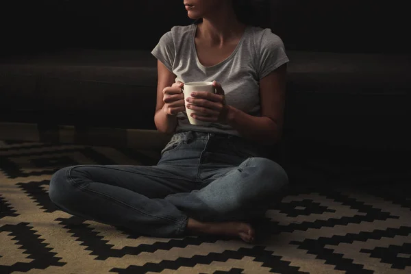 Cropped view of sad woman with coffee cup sitting on floor at home — Stock Photo
