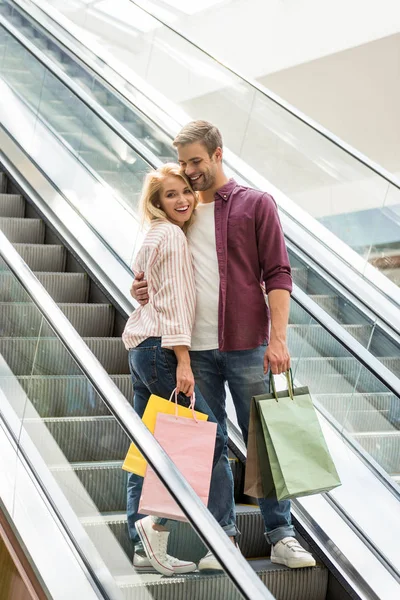 Guapo sonriente hombre con bolsas de compras abrazando novia en escalera mecánica en el centro comercial - foto de stock