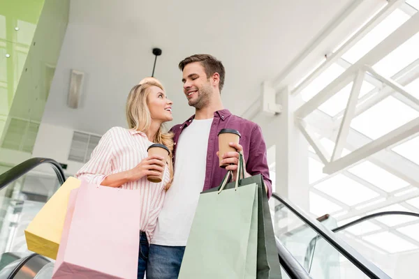 Vista de ángulo bajo de la joven pareja de compradores con bolsas de papel y tazas de café en escaleras mecánicas en el centro comercial - foto de stock