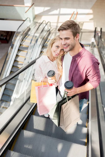 Vista de ángulo alto de la pareja con tazas de café desechables y bolsas de papel en la escalera mecánica en el centro comercial - foto de stock