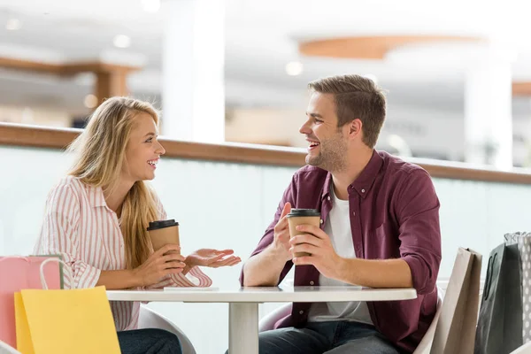 Sorrindo jovem casal com copos de papel de café sentado à mesa no café depois de fazer compras — Fotografia de Stock