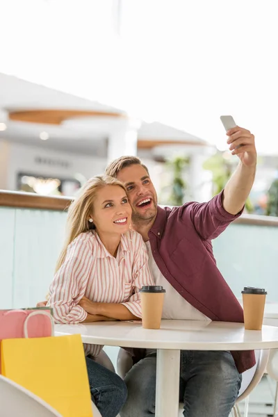 Pareja joven en la mesa con tazas de café tomando selfie en el teléfono inteligente en la cafetería - foto de stock