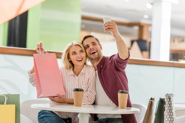 Hombre tomando selfie con novia mostrando bolsa de compras en la mesa con tazas de café desechables en la cafetería - foto de stock