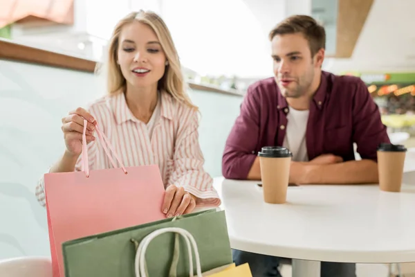 Selektiver Fokus der Frau, die Einkaufstasche vom Stuhl nimmt, während ihr Freund mit Kaffeetassen im Café am Tisch sitzt — Stockfoto