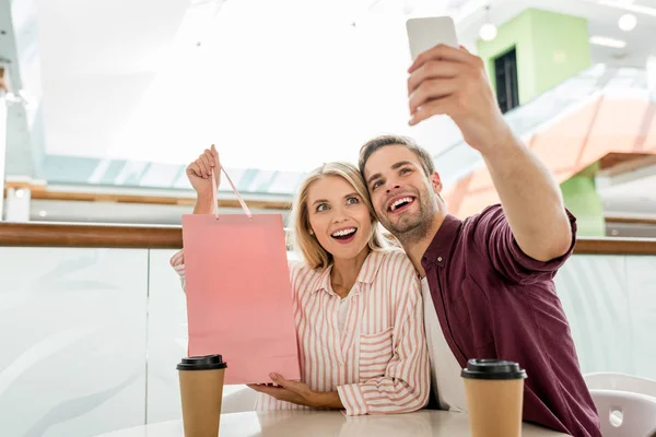 Young man taking selfie with girlfriend showing shopping bag at table with disposable cups of coffee in cafe — Stock Photo