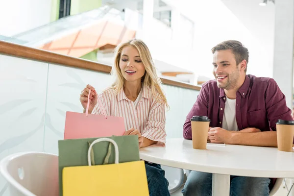 Attractive woman taking shopping bag from chair while her boyfriend sitting near at table with coffee cups in cafe — Stock Photo