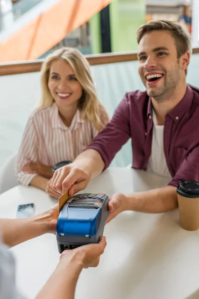 Jeune homme payant par carte de crédit sur le terminal tandis que sa petite amie assis près de la table avec des tasses à café dans le café — Photo de stock
