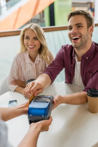 Junger Mann bezahlt mit Kreditkarte am Terminal, während seine Freundin in der Nähe mit Kaffeetassen im Café am Tisch sitzt — Stockfoto