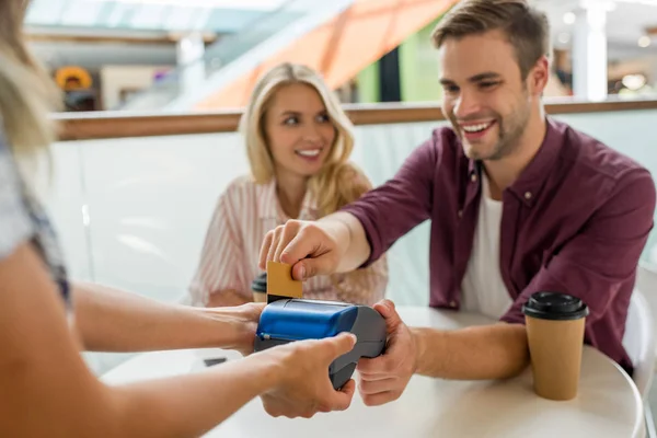 Foco seletivo jovem pagando com cartão de crédito no terminal, enquanto sua namorada sentada perto da mesa com xícaras de café no café — Fotografia de Stock