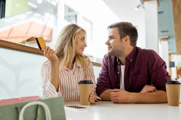 Casal com cartão de crédito e copos descartáveis de café olhando um para o outro à mesa no café — Fotografia de Stock