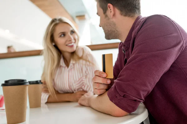 Enfoque selectivo de hombre joven con tarjeta de crédito hablando con su novia en la mesa con tazas de café en la cafetería - foto de stock