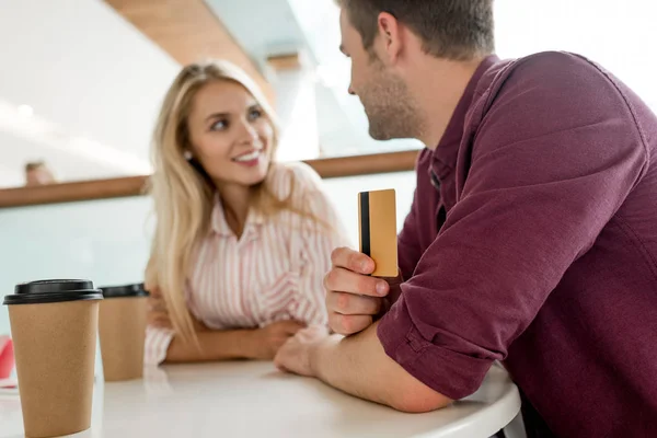 Foco seletivo do jovem com cartão de crédito conversando com a namorada à mesa com xícaras de café no café — Fotografia de Stock