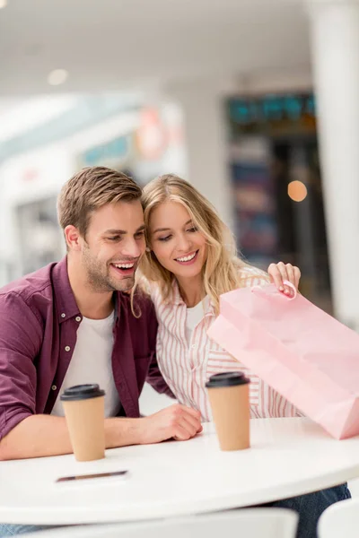 Selective focus of young woman looking into shopping bag with boyfriend at table with coffee cups in cafe — Stock Photo