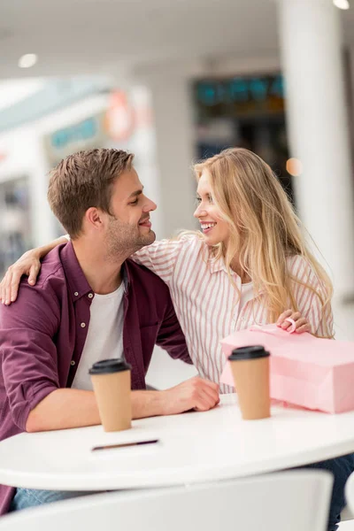 Sonriente pareja con bolsa de compras mirándose a la mesa con tazas desechables en la cafetería - foto de stock
