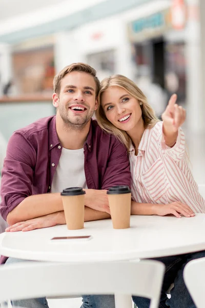 Attractive young woman pointing by finger to smiling boyfriend at table with disposable cups of coffee in cafe — Stock Photo