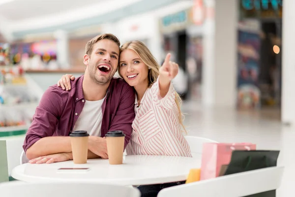 Enfoque selectivo de mujer joven atractiva señalando con el dedo a su novio sonriente en la mesa con tazas de café desechables en la cafetería - foto de stock