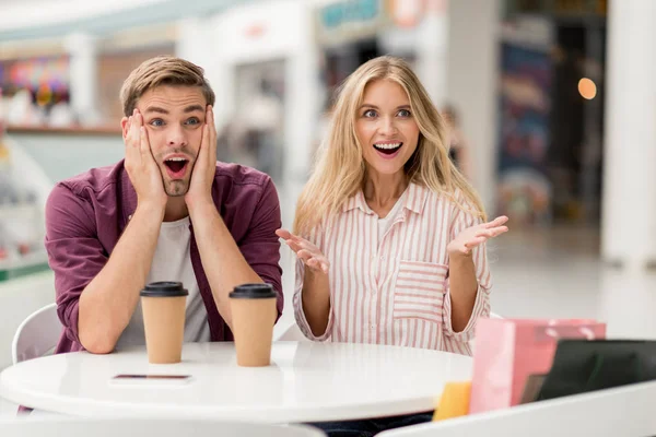 Sorprendida jovencita haciendo gestos con las manos mientras su impactado novio se sentaba cerca de la mesa con tazas de café en la cafetería - foto de stock