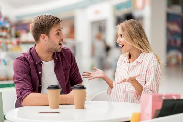 Shocked young woman gesturing by hands and looking at boyfriend while he sitting near at table with coffee cups in cafe — Stock Photo