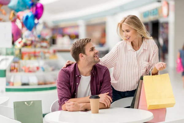 Giovane donna sorridente con borse della spesa in piedi vicino al fidanzato a tavola con tazza di carta di caffè nel caffè — Foto stock