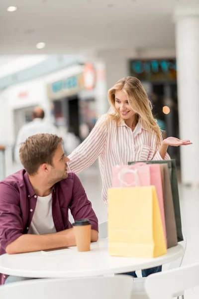 Bella giovane donna con borse della spesa gesticolando a mano e in piedi vicino al fidanzato a tavola con tazza di carta di caffè nel caffè — Foto stock