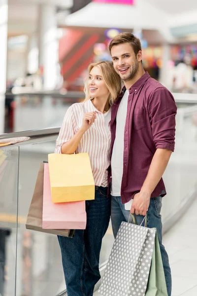 Молодая улыбающаяся пара с сумками и смартфоном в торговом центре — стоковое фото