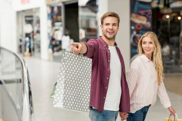 Selective focus of smiling young man with shopping bags pointing to girlfriend walking near at shopping mall — Stock Photo