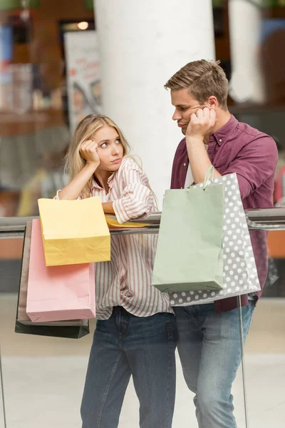 Chateado jovem casal de compradores com sacos de papel olhando uns para os outros no shopping — Fotografia de Stock