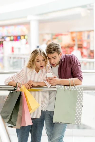 Шокированная парочка покупателей с бумажными пакетами, смотрящих на смартфон в торговом центре — стоковое фото