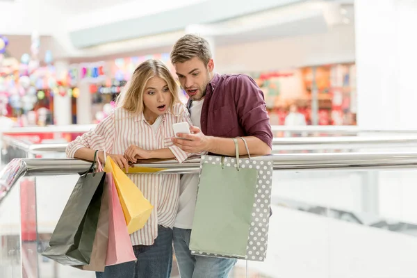 Joven pareja sorprendida con bolsas de papel mirando el teléfono inteligente en el centro comercial - foto de stock