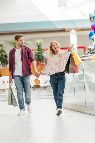 Красивая молодая женщина с сумками для покупок указывает на парня, идущего рядом с торговым центром — стоковое фото