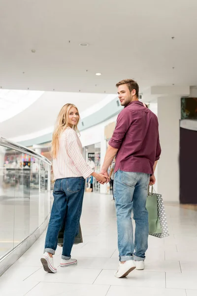 Sonriente pareja de compradores con bolsas de papel caminando en el centro comercial - foto de stock