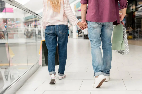 Imagen recortada de un elegante par de compradores con bolsas de papel cogidas de la mano y caminando en el centro comercial - foto de stock
