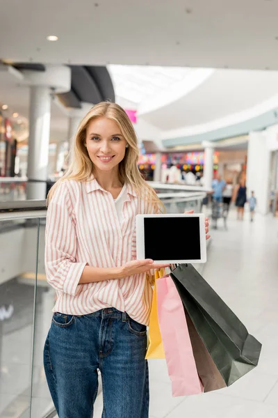 Приваблива усміхнена жінка з сумками, що показують цифровий планшет з порожнім екраном в торговому центрі — стокове фото