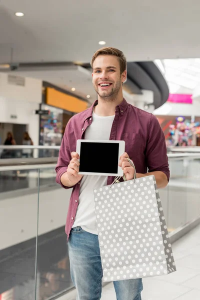 Jeune homme beau avec sac à provisions montrant tablette numérique avec écran blanc au centre commercial — Photo de stock