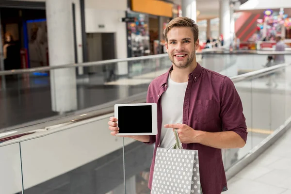 Joven hombre guapo con bolsa de compras apuntando con el dedo a la tableta digital con pantalla en blanco en el centro comercial - foto de stock