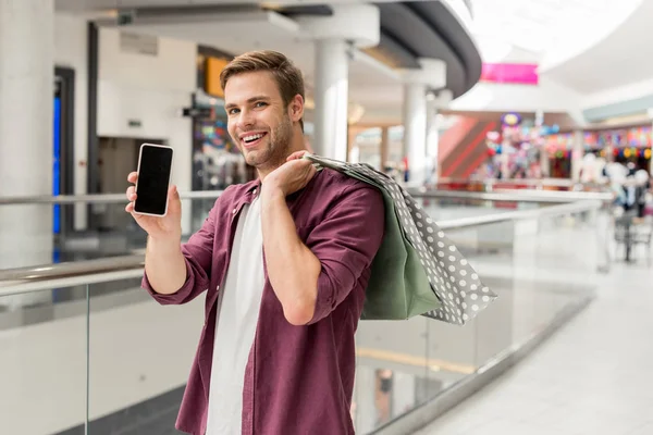 Giovane uomo sorridente con borse della spesa che mostra smartphone con schermo bianco al centro commerciale — Foto stock
