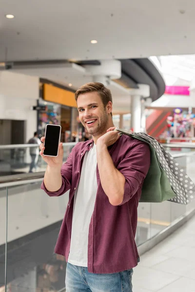 Jovem feliz com sacos de compras mostrando smartphone com tela em branco no shopping — Fotografia de Stock