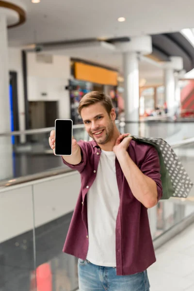 Junger Mann mit Einkaufstüten im Fokus zeigt Smartphone mit leerem Bildschirm in Einkaufszentrum — Stockfoto