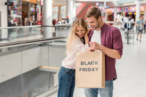 Jeune couple d'acheteurs regardant vers le bas et tenant sac en papier avec lettrage vendredi noir au centre commercial — Photo de stock