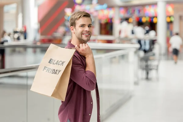 Giovane uomo sorridente in possesso di carta con lettering venerdì nero e guardando la fotocamera al centro commerciale — Foto stock