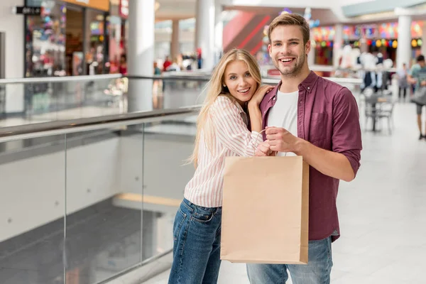 Glückliches junges Paar mit Papiertüte blickt in Kamera in Einkaufszentrum — Stockfoto