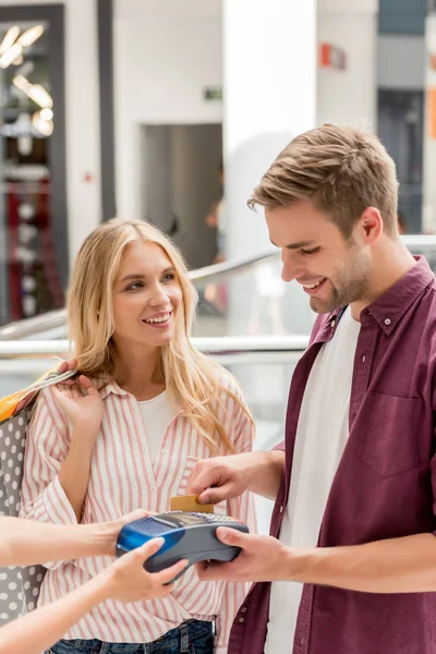 Улыбающийся мужчина платит кредитной картой в терминале, в то время как его девушка стоит рядом с мешками для покупок в торговом центре — стоковое фото
