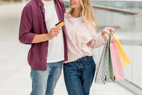 Visão parcial do homem mostrando cartão de crédito, enquanto sua namorada de pé perto com sacos de compras no shopping — Fotografia de Stock