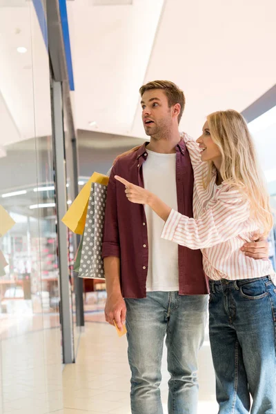 Emotional woman with shopping bags pointing by finger at shopwindow to shocked boyfriend at mall — Stock Photo