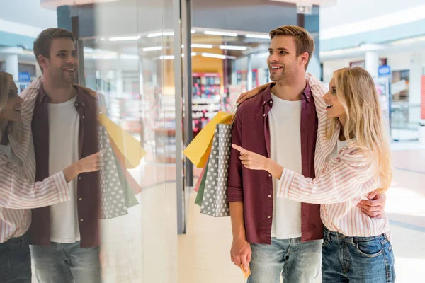 Young woman with shopping bags pointing by finger at shopwindow to boyfriend at mall — Stock Photo