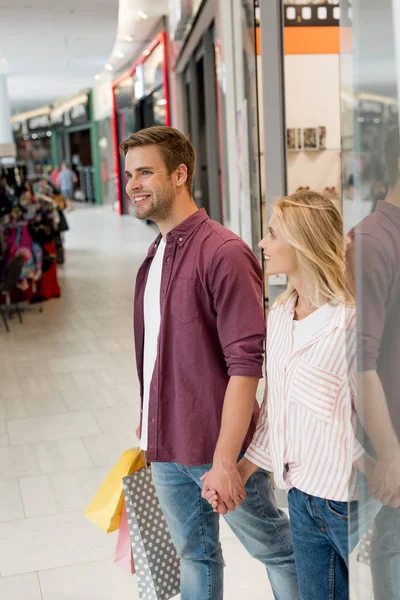 Smiling young couple with paper bags walking out from store at shopping mall — Stock Photo